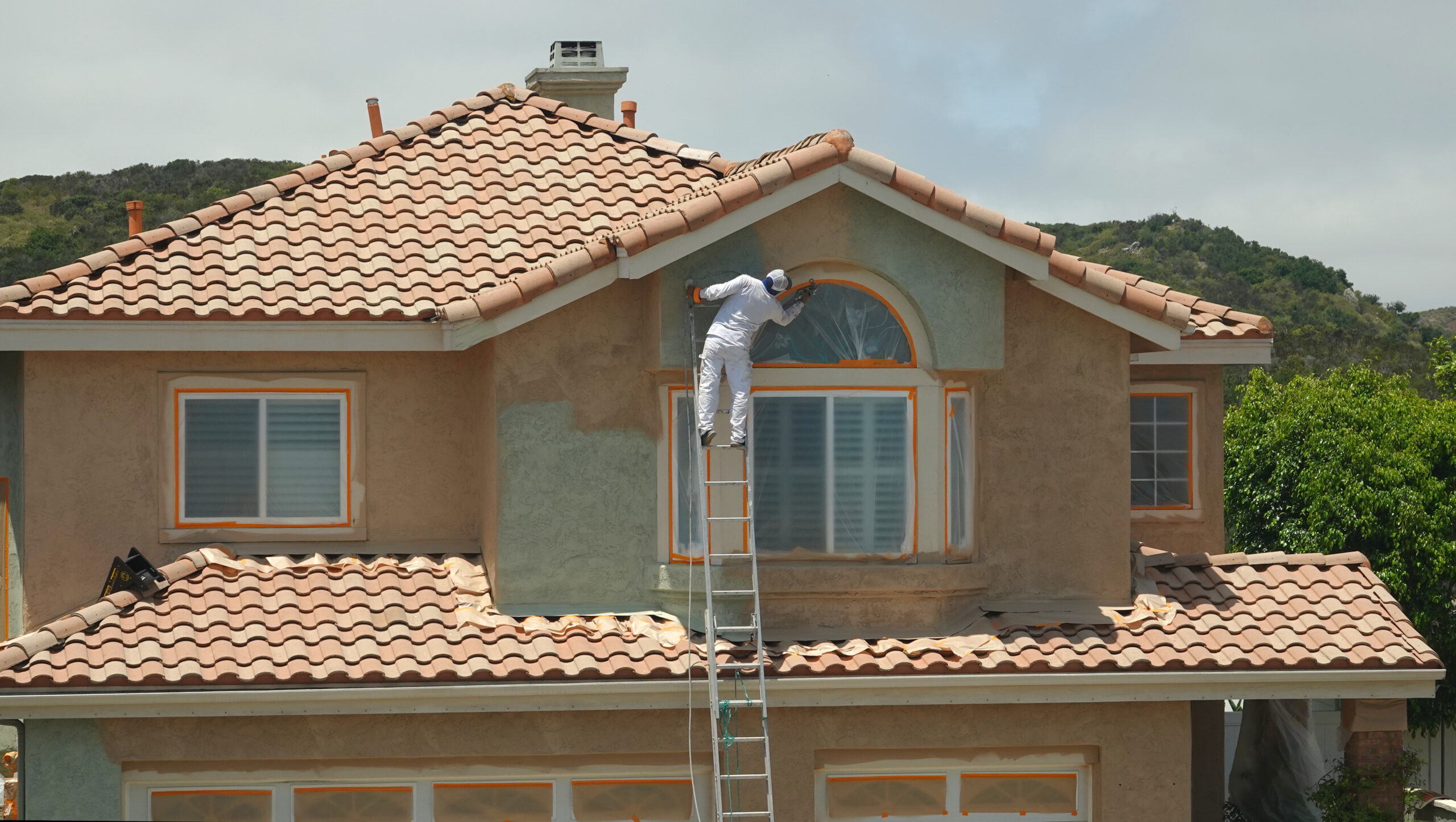 Real estate investor on a ladder painting the exterior of a two-story house during renovation—capturing the rehab phase of the BRRRR strategy in action.